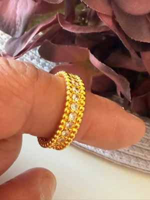 Guldfärgad ring i rostfritt stål med stenar - Snygg guldfärgad ring i rostfritt stål med glittrande stenar runt hela bandet. Ringen har en detaljrik, pärlad kant som ger en lyxig känsla och extra bling. Perfekt för dig som vill sticka ut med accessoarerna.storlek 16.5mm