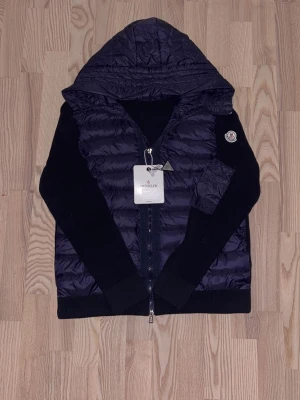 Mörkblå cardiganjacka från Moncler - Snygg mörkblå cardiganjacka från Moncler med stickade ärmar och quiltad kropp. Jackan har ett Moncler-märke på ärmen och innerfoder med serietryck. Perfekt för dig som vill ha en exklusiv och trendig look.