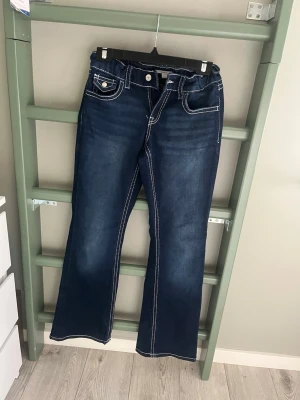 Mörkblå bootcut jeans med kontrastsömmar - Säljer ett par mörkblå bootcut jeans med tydliga vita kontrastsömmar och fem fickor. Jeansen har knapp- och dragkedjestängning samt dekorativa ficklock bak med knappdetaljer. Klassisk denimlook som passar till det mesta.