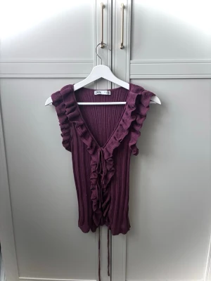 Jätte fin lila Zara topp - Supersnygg lila ribbad topp i S med volangdetaljer längs v-ringningen och framtill. Ärmlös modell med knytband framtill som ger en extra touch. Perfekt att styla med jeans eller kjol för en trendig look. 