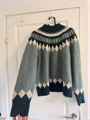 Grön stickad tröja från ONLY - Mysig stickad tröja från ONLY i mörkgrönt med beige och vitt mönster. Tröjan har högre krage, långa ärmar och ribbstickade muddar. Perfekt för kalla dagar och har en snygg retroinspirerad look med grafiskt mönster🪻