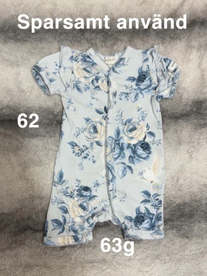 Blommig baby-pyjamas kort ärm - Supersöt baby-pyjamas i ljusblått med blå och vita blommor. Kortärmad och kortbent modell med tryckknappar framtill som gör den enkel att ta på och av. Perfekt för varma nätter och mjukt tyg som känns skönt mot huden.🌸 Sparsamt använd. Storlek 62