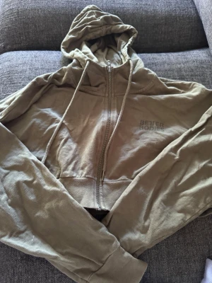 Beige cropped hoodie Better Bodies - Snygg grön cropped hoodie från Better Bodies med dragkedja framtill och huva. Diskret logga på bröstet och stor BB-logga på ryggen. Tillverkad i mjukt material som passar perfekt till träning eller chill. Långa ärmar och avslappnad passform.