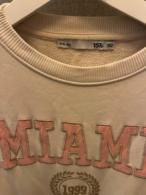 Beige sweatshirt med MIAMI-tryck - Säljer en beige sweatshirt med texten MIAMI i rosa och vitt på bröstet samt ett tryck med 1999 och lagerkrans under. Tröjan har rund halsringning, ribbade muddar och är gjord i mjukt sweatshirtmaterial. Perfekt för en avslappnad och trendig look.                       Ska strykas vid köp!!!