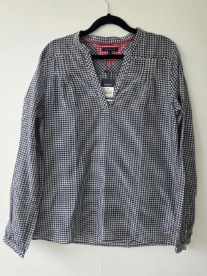 Rutig blus från Tommy Hilfiger - Snygg blus med prislappen på från Tommy Hilfiger i svartvitt rutigt mönster. Blusen har V-ringning, långa ärmar och en avslappnad passform. Tillverkad i ett lätt och mjukt tyg som känns skönt mot huden. Perfekt för en trendig och avslappnad look.