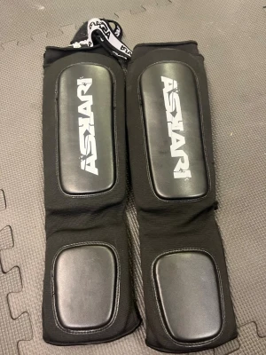 Svarta benskydd från Askari strl M - Säljer ett par svarta benskydd från Askari, perfekta för kampsport. De har vadderade skyddszoner på framsidan och vid foten, samt elastiska band för att hålla dem på plats. Materialet är en kombination av slitstark textil och konstläder.