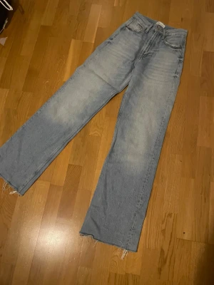 Ljusblå raka jeans med rå kant - Säljer ett par ljusblå jeans med rak passform och hög midja. Jeansen har klassiska fem fickor och råa, fransiga benslut för en avslappnad vibe. Tillverkade i denim med lätt tvättad look och slitningar på benen.
