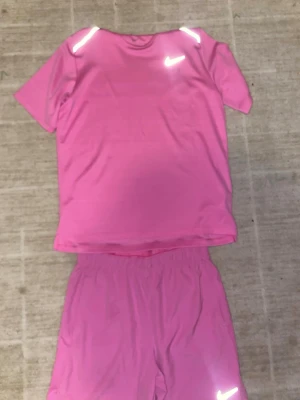 Rosa träningsset från Nike - Säljer ett rosa träningsset från Nike med t-shirt och shorts. Setet är gjort i ett lätt och snabbtorkande material, perfekt för träning. Både tröjan och shortsen har vita Nike-loggor och reflekterande detaljer. T-shirten har rund hals och shortsen har resår i midjan. Tröjan har storleken XS altaå XL i junior size och byxorna L size på junior så alltså en S. Inga deffekter andvänd 2-3 gånger men mycket fin skick. Priset är spikat