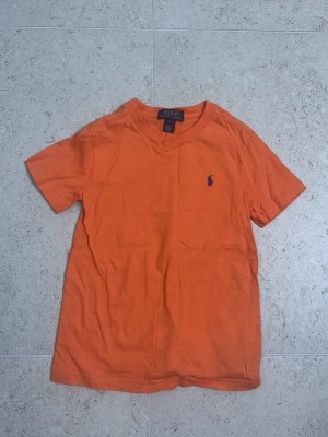 Orange t-shirt från Polo Ralph Lauren barn - Snygg orange t-shirt från Polo Ralph Lauren med klassisk v-ringning och den ikoniska lilla loggan broderad på bröstet. Tillverkad i mjuk bomull och har en normal passform med korta ärmar. Perfekt för en avslappnad och stilren look. Storlek för 6 åringar.