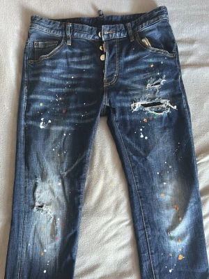 Blåa Dsquared2 jeans med färgstänk S: M48 - Säljer ett par blå jeans med slitna detaljer och färgstänk i olika färger. Jeansen har raka ben, klassisk femficksmodell och knappgylf. Perfekta för dig som gillar en kreativ och avslappnad stil.