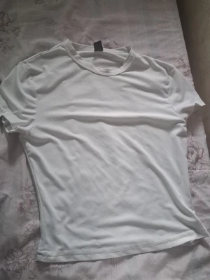 Vit basic t-shirt i bomull - Klassisk vit t-shirt med rund hals och korta ärmar. Modellen har en normal passform och är gjord i mjukt bomullsmaterial som känns skönt mot huden. Perfekt att styla till jeans eller kjol.
