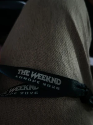The Weeknd Europe 2026 tygarmband - Säljer ett svart tygarmband med vit text 'THE WEEKND EUROPE 2026'. Armbandet har en vit plastdetalj och är perfekt som minne från konserten eller som accessoar för fans. Snygg och enkel design som passar till många stilar.