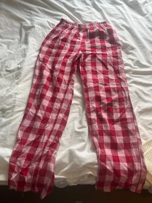 Rödrutiga pyjamasbyxor från H&M - Säljer ett par rödrutiga pyjamasbyxor från H&M i storlek 170. Byxorna har ett klassiskt rutigt mönster i rött och rosa, med resår i midjan för extra komfort. Perfekta för mysiga kvällar hemma.