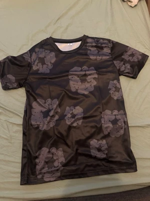 Svart t-shirt med grått camomönster - Cool svart t-shirt med grått camouflagemönster. Tillverkad i ett glansigt syntetmaterial som känns lätt och smidigt. Klassisk passform med rund hals och korta ärmar. Perfekt för en avslappnad streetwear-look.