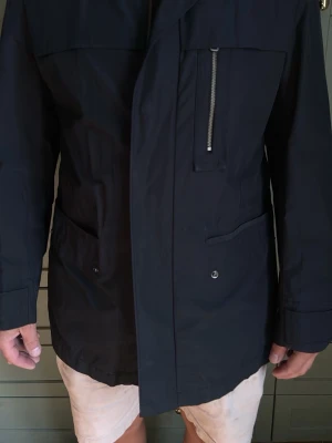 Mörkblå vindjacka med dragkedja - Snygg mörkblå vindjacka med rak passform och flera fickor, inklusive en bröstficka med dragkedja. Jackan har knappstängning på fickorna och justerbara ärmslut. Perfekt för blåsiga dagar och enkel att matcha med olika outfits.