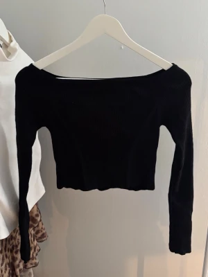 Svart ribbad offshoulder topp - Trendig svart ribbad topp med offshoulder-design och långa ärmar. Croppad modell som passar perfekt till höga jeans eller kjol. Mjuk och stretchig känsla för en bekväm passform.