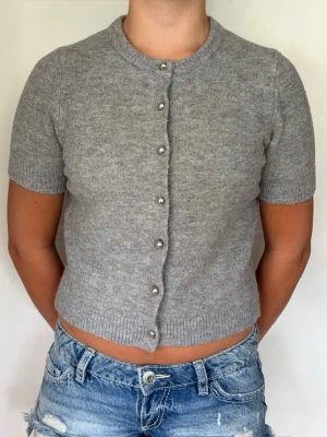 Grå kortärmad kofta från Zara - Kortärmad grå kofta från Zara med rund halsringning och dekorativa pärlknappar framtill. Koftan är stickad i mjukt material och har ribbade kanter vid ärmslut och nederkant. Perfekt att styla med jeans eller kjol.