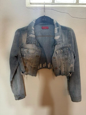 Cropped ljusblå jeansjacka - Säljer en trendig cropped jeansjacka i ljusblå denim med slitna detaljer och rå kant nertill. Jackan har två bröstfickor med knappar och klassisk krage. Perfekt för dig som vill ha en edgy och avslappnad stil.