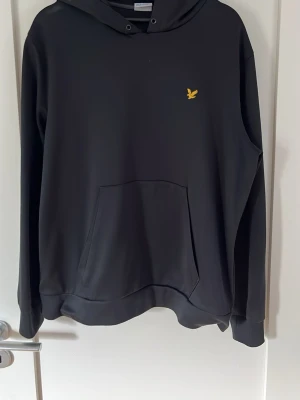 Svart hoodie med gul logga - Snygg svart hoodie med huva och känguruficka framtill. På bröstet sitter en liten gul broderad logga som ger en clean look. Hoodien har ribbade muddar vid ärmar och nederkant. Perfekt för dig som gillar enkel och stilren streetwear.