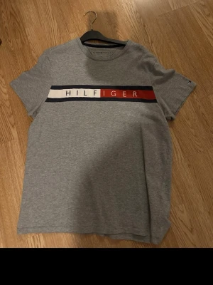 Grå Tommy Hilfiger t-shirt - Snygg grå t-shirt från Tommy Hilfiger med klassiskt tryck i vitt, blått och rött över bröstet. Rund halsringning och korta ärmar. Tillverkad i mjukt bomullsmaterial som känns skönt mot huden. Perfekt för en avslappnad och stilren look.