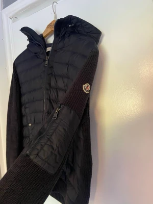 moncler cardigan  - Snygg svart pufferjacka från Moncler med stickade ärmar och huva. Jackan har dragkedja framtill, fickor med knappar och en extra ficka med dragkedja på ärmen. Moncler-logga på vänster ärm. Perfekt för kalla vinterdagar. storelk 2 = S-M