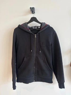 Svart zip hoodie från Burberry XS - Snygg svart hoodie från Burberry med dragkedja, huva med grått och rött foder samt fickor framtill. Klassisk Burberry-logga på bröstet. Köpt i burberry butik i Italien av mig, kvitto finns. Bra skick 8/10. Storlek xs, sitter som en S