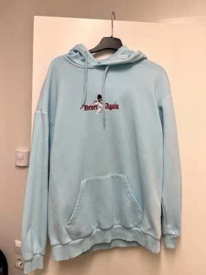 Ljusblå hoodie med tryck och huva - Säljer en ljusblå hoodie med huva och dragsnören. På bröstet finns ett tryck med texten 'Never Again' och en liten illustration. Hoodien har en stor magficka och ribbade muddar vid ärmar och nederkant. Perfekt för en chill och avslappnad stil.