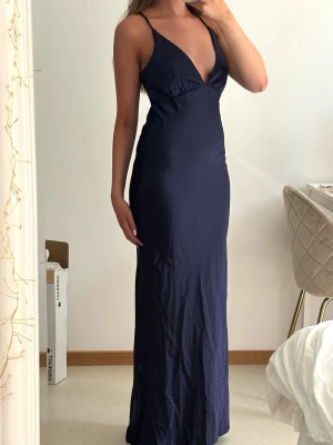 Marinblå v-ringad balklänning med smala axelband  - Snygg marinblå långklänning med smala axelband och djup v-ringning. Klänningen har en figurnära passform och är ärmlös, vilket ger en elegant och stilren look. Perfekt för bal, bröllop eller finare sittningar. 