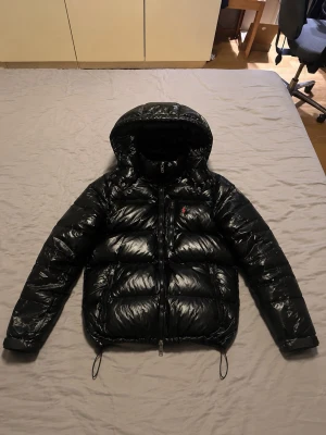 Svart glansig dunjacka med huva - Säljer en svart Ralph lauren jacka som är i nytt skick och är riktigt fet, jag säljer den eftersom jag har en annan jacka som jag föredrar. Jackan har endast använts ett fåtal gånger då jag har en annan jacka. Köpte den för 4999kr på rabatt säljer den nu för 1649kr då jag vill få något för den istället för att den bara ska ligga hemma.