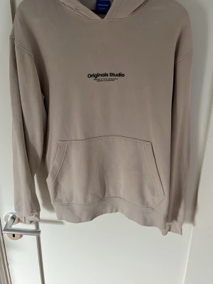 Beige hoodie Originals Studio - Säljer en beige hoodie från Originals Studio med svart text på bröstet. Hoodien har huva, känguruficka framtill och ribbade muddar vid ärmar och nederkant. Perfekt för en chill och avslappnad stil.