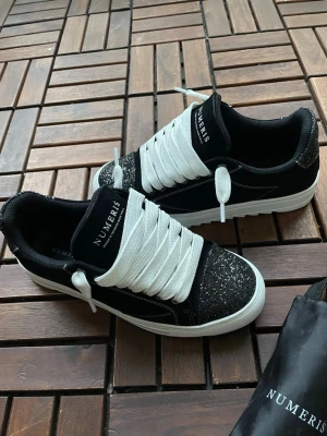Numeris - Snygga svarta sneakers från Numeris med vit tjock sula och breda vita skosnören. Tåpartiet är täckt av svart glitter som ger extra attityd. Ovandelen är i textil och mockaimitation, och loggan syns tydligt på plösen. Perfekta för dig som vill sticka ut.