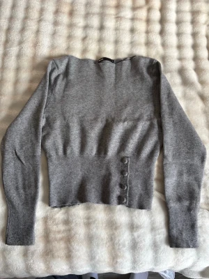 Grå stickad tröja med knappar - Snygg grå stickad tröja med bred ribbad midja och fyra dekorativa knappar på sidan. Tröjan har lång ärm och en enkel, stilren design som passar perfekt till jeans eller kjol. Mjuk och skön kvalitet som håller dig varm under kyliga dagar.