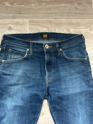 Mörkblå jeans från Lee - Snygga mörkblå jeans från Lee med klassisk femficksmodell och kontrastsömmar. Jeansen har en rak passform och detaljer i guld, samt ett brunt läderpatch bak i midjan. Perfekta för en avslappnad och trendig look.