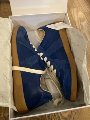 Blå sneakers Maison Martin Margiela - Snygga blå sneakers från Maison Martin Margiela med ovandel i mocka och läder, vita snören och klassisk gummisula i brunt. Skorna har en stilren design med paneler och låg profil som passar till många olika outfits.