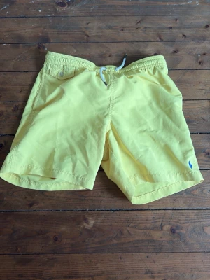 Gula badbyxor från Polo Ralph Lauren - Snygga gula badbyxor från Polo Ralph Lauren med klassisk blå logga broderad på benet. Modellen har elastisk midja med dragsko och sidofickor. Perfekta för stranden eller poolen, tillverkade i snabbtorkande material.