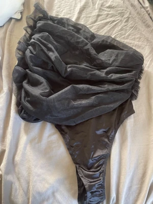 Svart body med volangdetalj - Unik svart body med stor volangdetalj över ena axeln. Bodyn har en asymmetrisk design och är tillverkad i glansigt syntetmaterial. 