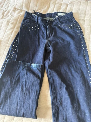 Mörkblå jeans med nitar från Bershka - Säljer ett par mörkblå jeans från Bershka med raka ben och coola silverfärgade nitar längs fickor och sidor. Jeansen har en patchad detalj på ena benet och klassisk femficksmodell. Perfekta för dig som vill sticka ut med din stil!