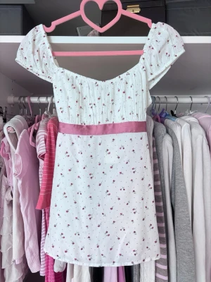 Blommig vit blus med rosett - Supersöt vit blus med små rosa blommor och en bred rosa rosett i midjan. Blusen har offshoulder-korta puffärmar och smock upptill för en bekväm passform. Perfekt för dig som gillar romantisk och feminin stil.