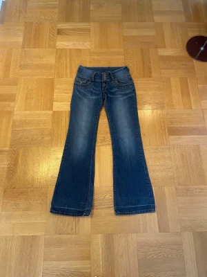 Blå bootcut jeans med låg midja - Säljer ett par blå bootcut jeans med låg midja och fyra knappar i gylfen. Jeansen har slitningar framtill, klassiska fem fickor och kontrastsömmar. Perfekta till hösten och för dig som gillar 2000s stil. Mått: Midja: 33,5 cm Gren: 20 cm Övre lår: 21 cm Innerbenslängd: 80 cm