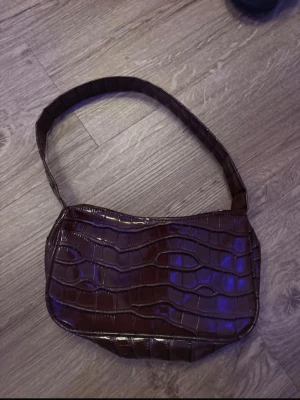 Brun axelväska i krokodilimitation - Snygg brun axelväska med croco-mönster och glansig finish. Väskan har en enkel, rektangulär form och bred axelrem. Perfekt för dig som gillar trendiga accessoarer med lite edge.
