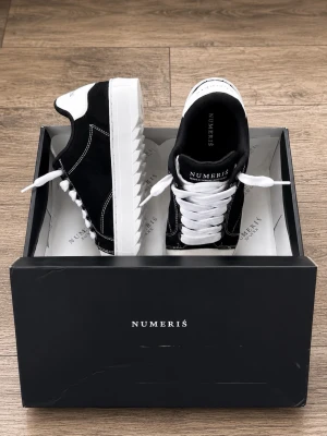  Numeris - Stilrena sneakers från Numeris i svart mocka och vitt läder med vita kontrastsömmar och tjock vit sula. Skorna har vita skosnören och en låg profil som ger en clean look. Perfekta för dig som gillar klassisk streetstyle.