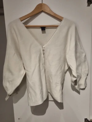 Vit kofta med v-ringning från Lindex (Har en fläck, bild 4) - Stilren vit kofta från Lindex med v-ringning och tre knappar framtill. Koftan har lite vidare ärmar och är gjord i ett mjukt, stickat material. Perfekt att slänga över en topp för en enkel och snygg look.