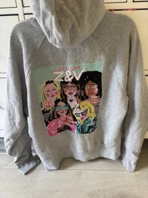 Grå hoodie med broderad Z&V design - Säljer en grå hoodie från Zadig & Voltaire med en stor broderad färgglad design baktill som föreställer fem personer och texten Z&V. Hoodien har huva med vita snören och en klassisk magficka. Perfekt för dig som gillar unika prints och streetstyle. Skulle säga att den passar S/M (overzised S i storlel)