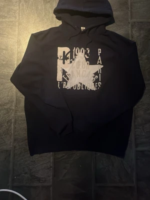 Marinblå hoodie från Zandro studio - Snygg marinblåhoodie med stor vit stjärna och texttryck på framsidan. Klassisk modell med huva och långa ärmar. Perfekt för en avslappnad och cool stil. Materialet känns mjukt och bekvämt, passar perfekt till jeans eller joggers.