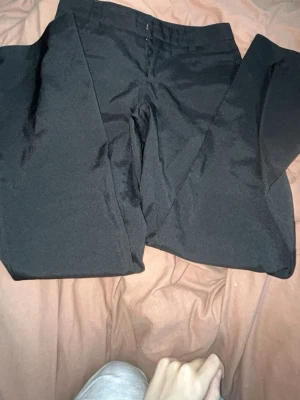 Svarta kostymbyxor från Gina Tricot - Stilrena svarta kostymbyxor från Gina Tricot med klassisk rak passform och hällor i midjan. Perfekta för en dressad look och enkla att matcha med olika toppar. Materialet är mjukt och har en lätt stretch för extra komfort.