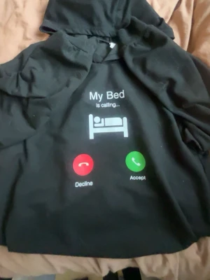 Svart hoodie med tryck från SHEIN - Svart hoodie från SHEIN med huva och ett roligt tryck på bröstet: 'My Bed is calling...' tillsammans med en sängikon och knappar för att acceptera eller neka samtalet. Perfekt för dig som gillar chill vibes och humoristiska plagg.