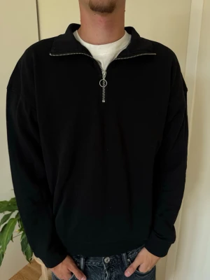 Svart halvzip sweatshirt - En svart sweatshirt med halvzip och hög krage. Tröjan har långa ärmar och en avslappnad passform, perfekt att slänga på sig över en t-shirt. Materialet ser ut att vara mjukt och bekvämt, troligen bomull eller bomullsblandning.