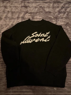 Svart stickad Saint Laurent tröja - Säljer en svart stickad tröja från Saint Laurent med vit logotyp i handskriven stil på bröstet. Tröjan har rund halsringning, långa ärmar och ribbade muddar vid ärmslut och nederkant. Perfekt för dig som gillar exklusiva och stilrena plagg.