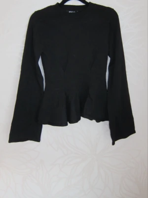 Svart långärmad topp med peplum - Snygg svart långärmad topp med peplumdetalj i midjan. Toppen har en enkel rund halsringning och är tillverkad i ett mjukt material som ger en bekväm passform. Perfekt att styla med jeans eller kjol för en trendig look.