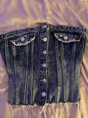 Jeans korsett-topp med knappar - Snygg jeans tub topp med knappar framtill och två fickor upptill. Slitdetaljer och råa kanter nedtill ger en edgy look. Smockad rygg för skön passform. Köpt på plick och nästan aldrig använd! 